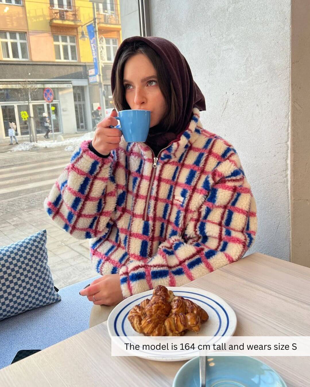 Model wearing a stylish oversized 100% merino wool jacket in pink and cobalt, sipping tea with croissants.